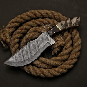 May include: A handcrafted Damascus steel knife with a textured blade and a dark wooden handle with gold accents. The knife is displayed on a coiled brown rope, showcasing its intricate design and craftsmanship. The blade has a unique pattern.