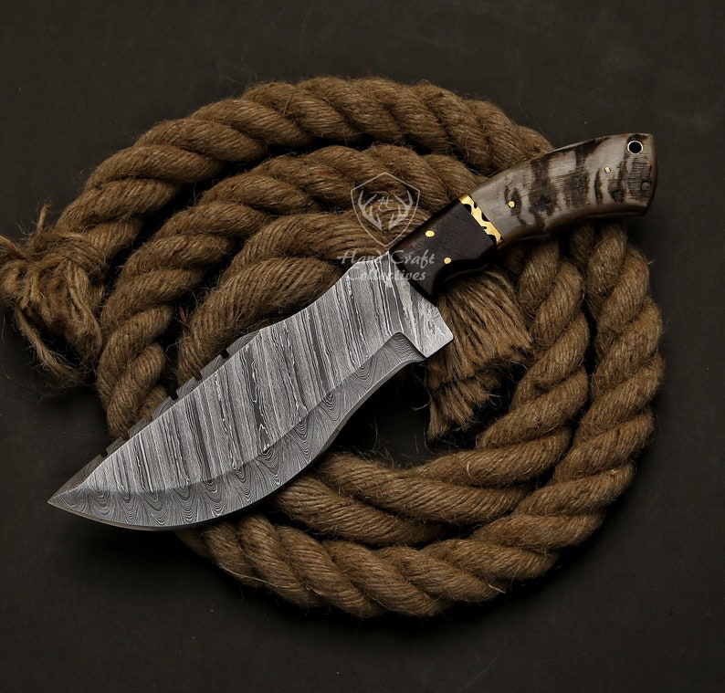 May include: A handcrafted Damascus steel knife with a textured blade and a dark wooden handle with gold accents. The knife is displayed on a coiled brown rope, showcasing its intricate design and craftsmanship. The blade has a unique pattern.