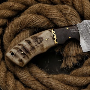 May include: Close-up of a handcrafted knife with a patterned blade and a handle made of bone and dark wood. The handle is accented with brass details. The knife rests on a bed of thick, brown rope.
