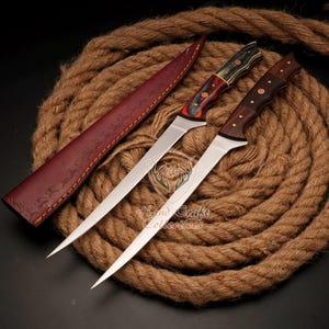 May include: Two long, thin, silver-colored knives with wooden handles. The handles are red, black, and brown. The knives are on a brown rope background. The knives are in a leather sheath.