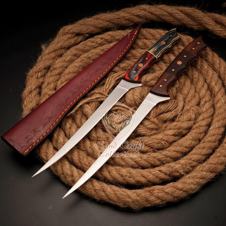 May include: Two long, thin, silver-colored knives with wooden handles. The handles are red, black, and brown. The knives are on a brown rope background. The knives are in a leather sheath.