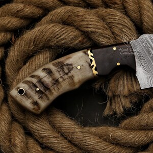 May include: Close-up of a handcrafted knife with a patterned blade and a handle made of bone and dark wood. The handle is accented with brass details. The knife rests on a coil of thick, brown rope.