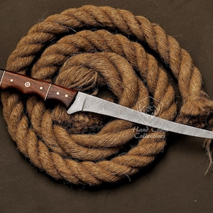 May include: A hand-crafted, Damascus steel knife with a dark wood handle. The blade has a wavy pattern and is very sharp. The knife is resting on a coil of brown rope.