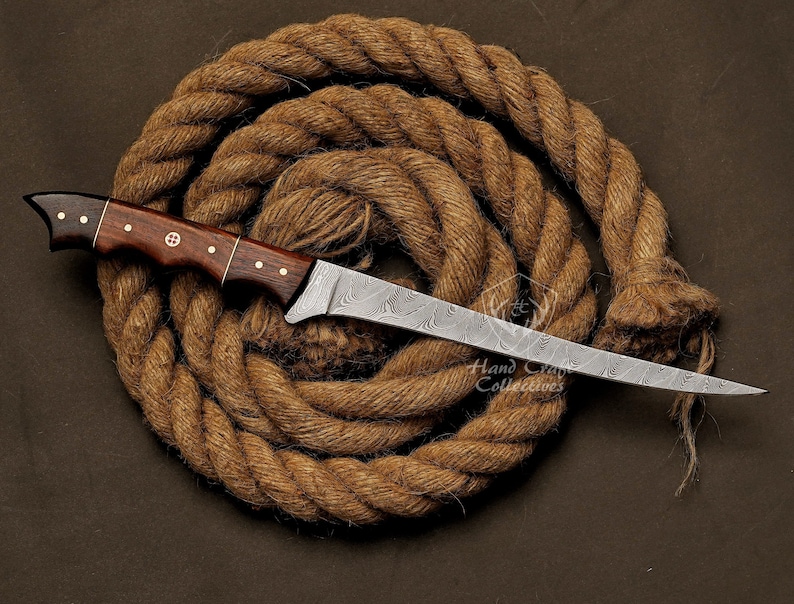 May include: A hand-crafted, Damascus steel knife with a dark wood handle. The blade has a wavy pattern and is very sharp. The knife is resting on a coil of brown rope.