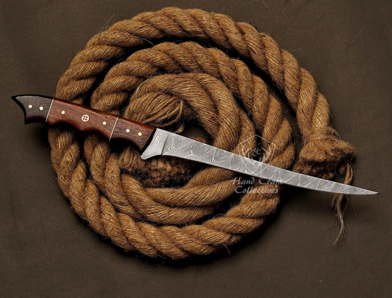 May include: A hand-crafted, Damascus steel knife with a dark wood handle. The blade has a wavy pattern and is very sharp. The knife is resting on a coil of brown rope.