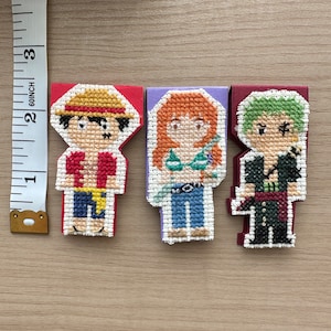 May include: Three embroidered felt bookmarks featuring characters from the anime One Piece. The bookmarks are red, purple, and green. The characters are Luffy, Nami, and Zoro.