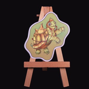 May include: A green and brown turtle with a red saddle riding on top of another green and brown turtle. The turtles are on a small wooden easel.