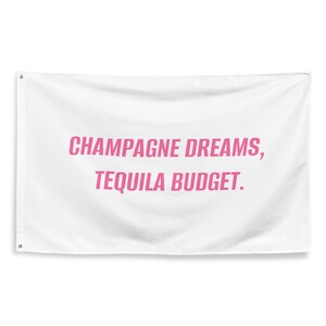 May include: White rectangular wall flag with the text "CHAMPAGNE DREAMS, TEQUILA BUDGET" in pink lettering. The flag has grommets in each corner for hanging. The flag is made of a soft, slightly textured fabric.
