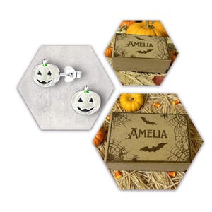 May include: Silver pumpkin earrings with black and white detailing. The earrings are displayed on a grey hexagonal surface. Two brown boxes with the name "AMELIA" are also shown, decorated with spiderwebs and bats, suggesting a Halloween theme.