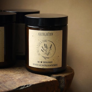 Könnte beinhalten: Ein dunkelbraunes Kerzenglas mit schwarzem Deckel. Das Etikett trägt die Aufschrift "Kerzengarten" und "Lavendel & Zeder & Tanne" mit einer botanischen Illustration. Der Text "Mit handgemacht Duftkerze aus natürlichem Rapswachs" ist ebenfalls sichtbar. Die Kerze steht auf einer Holzoberfläche.