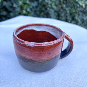 May include: Handmade ceramic mug with a unique color gradient. The mug transitions from a dark blue base to a reddish-brown middle, and a white rim. The handle is dark blue. This handcrafted mug is perfect for coffee or tea.