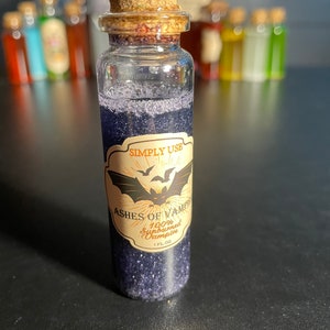 May include: A small glass bottle with a cork stopper filled with a dark purple liquid and glitter. The bottle has a label that reads "Simply Use Ashes of Vampire 100% Sunburned Vampire 1 fl oz".
