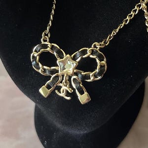 May include: Gold-tone chain necklace with a black leather and gold bow pendant. The bow features a clear star-shaped crystal and a small gold logo charm. The necklace is displayed on a black velvet surface.