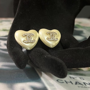 May include: A pair of heart-shaped earrings in a cream-colored finish, each featuring a gold-tone interlocking CC logo embellished with clear crystals centered on the heart. The earrings have a smooth, glossy surface and a rounded, puffed design.
