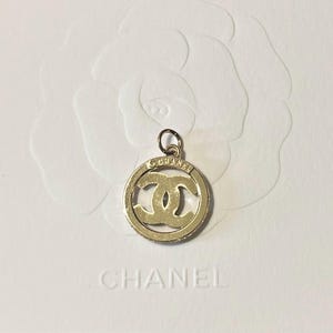 以下が含まれることがあります： シャネルのロゴ、二重に絡み合ったCのデザインが施されたゴールドカラーのペンダント。ペンダントは円形で、ブランド名が縁に刻まれています。背景は白い花柄で、ブランド名が描かれています。