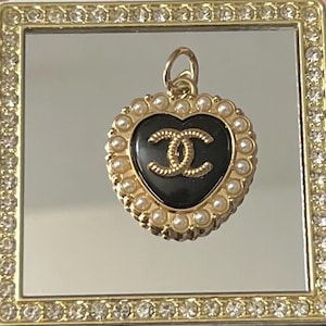 May include: A heart-shaped pendant with a black center and a gold-colored double C logo. The pendant is surrounded by small, white pearls and a gold-colored frame. The pendant is displayed on a mirrored surface.