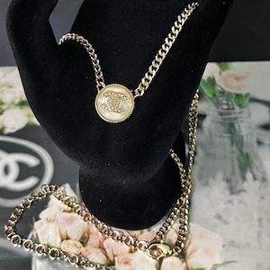 May include: A gold-tone chain necklace featuring a circular pendant with a white, iridescent center and a gold interlocking CC logo. The chain includes a lobster clasp closure and a small dangling CC charm at the end, showcasing a classic link design.