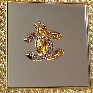 May include: A gold-colored pendant with a double C design, embellished with small, clear crystals. The pendant is set against a mirrored background and framed by a gold border encrusted with clear crystals. The pendant has a bee-like design.