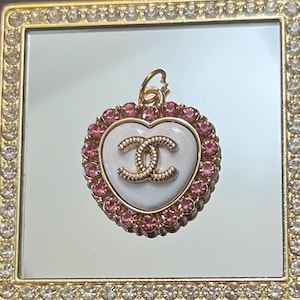 May include: Gold-tone heart-shaped pendant with a white centre featuring a gold logo. The pendant is surrounded by pink crystals and has a small loop at the top for attachment. The pendant is displayed on a mirrored surface.