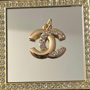 May include: A gold-colored pendant with a double-C design, encrusted with small, clear crystals. The pendant hangs from a gold loop. The pendant is set against a mirrored surface within a square, gold-framed compact, also adorned with crystals.