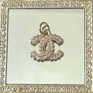 May include: A gold-toned metal pendant featuring the interlocking CC logo design, embellished with small white faux pearls and clear sparkling crystals. The pendant includes a small circular jump ring at the top for attachment to a chain or cord.