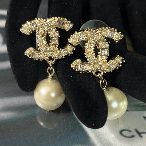 May include: A pair of gold-tone earrings featuring the interlocking CC logo design. Each logo is embellished with small white pearls and clear square-cut crystals. A single, larger round white faux pearl hangs from the base of each logo.