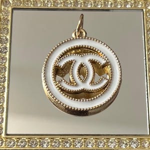 May include: A gold-colored pendant with a white enamel border and a gold double-C logo. The pendant is round and has a small loop at the top for a chain. The pendant is set against a gold-framed mirror.