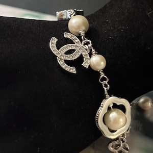 May include: Silver-toned bracelet featuring a sparkling logo charm and a floral-shaped pendant, both adorned with clear crystals. The bracelet includes several faux pearls, adding a touch of elegance to the design. A classic piece of jewelry.