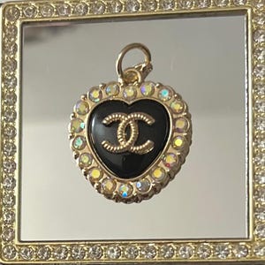 May include: A gold-toned heart-shaped pendant with a black center featuring a gold double-C logo. The pendant is surrounded by iridescent crystals and has a small loop at the top for hanging.