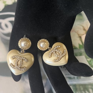 May include: A pair of gold-tone earrings featuring a round pearl stud connected to a heart-shaped pendant. The heart is a smooth cream-colored material with a central crystal-encrusted interlocking CC logo. The stud is bordered by small clear crystals.