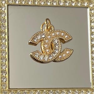 May include: Gold-toned charm with a double-C design, encrusted with clear crystals and small pearls. The charm is suspended from a small loop. The charm is set against a mirrored surface within a gold-framed square, also adorned with crystals.