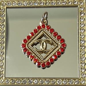 May include: A gold-toned pendant with a diamond-shaped frame, adorned with red gemstones. The center features a double-C logo. The pendant is set within a gold-framed mirror, embellished with clear crystals, creating a luxurious aesthetic.