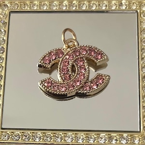 May include: A gold-toned pendant in the shape of interlocking C's, encrusted with pink rhinestones. The pendant has a small gold loop at the top for hanging. The pendant is displayed on a mirrored surface within a gold-framed box.