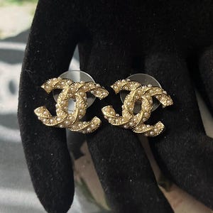 May include: A pair of gold-toned stud earrings in a double-C design, encrusted with small, round, off-white pearls. The earrings are displayed against a dark background, highlighting their intricate details and elegant style.