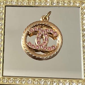 May include: A gold-toned pendant with a double-C design encrusted with pink gemstones. The pendant is encircled by a twisted rope-like border. The pendant is displayed on a reflective surface within a gold-framed square.