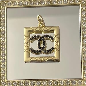 May include: Gold-toned pendant with a square frame, featuring a double-C logo embellished with black stones. The frame has a quilted pattern and is surrounded by a border of clear crystals. The pendant is designed for jewelry.