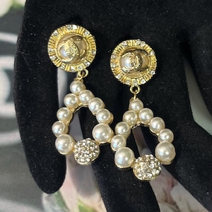 May include: A pair of gold-tone drop earrings featuring a circular stud with a central CC logo surrounded by clear crystals. Below, a teardrop-shaped frame is adorned with white faux pearls and a single cluster of clear crystals at the base.