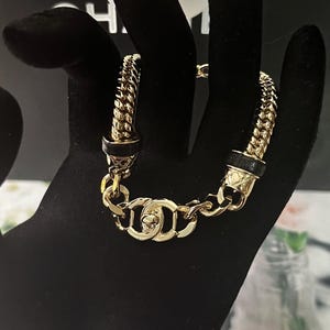 May include: Gold-tone chain bracelet with a woven design and black leather accents. The bracelet features a series of interlocking links and a decorative clasp. A luxury jewelry item.