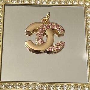 May include: A gold-colored charm in the shape of interlocking letters, embellished with pink rhinestones. The charm is suspended from a small loop and is set against a reflective surface within a decorative, jewel-encrusted frame.