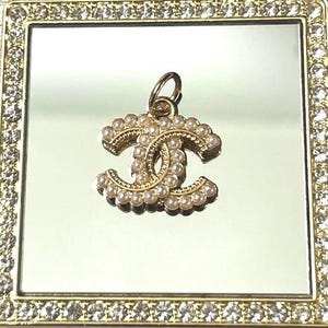 May include: A gold-toned metal pendant featuring the interlocking CC logo design, embellished with small white faux pearls along the surface. The pendant includes a circular jump ring at the top for attachment and is centered on a square mirror.