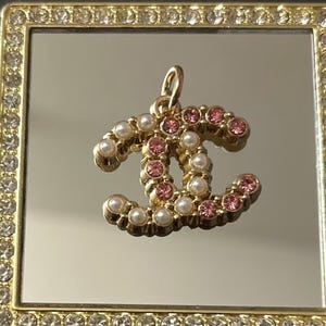 May include: Gold-toned pendant in a double-C design, adorned with pink and white gemstones. The pendant is set against a mirrored surface within a square, gold-framed border embellished with clear crystals. The overall aesthetic is elegant and luxurious.