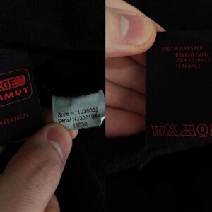 Puede incluir: Primer plano de tela negra con varias etiquetas. Una etiqueta presenta el texto rojo "LARGE MAMMUT" y "MADE IN PORTUGAL". Otra etiqueta muestra "Style N. 1030031" y "Serial N.:3001044". Una tercera tiene instrucciones de lavado y el texto "100% POLYESTER".