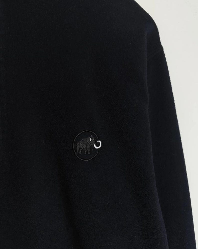 Puede incluir: Chaqueta polar negra con un parche redondo que presenta un elefante negro y un colmillo blanco curvado. La chaqueta tiene un aspecto suave y texturizado y un dise&ntilde;o sencillo. El parche est&aacute; bordado con finos detalles.