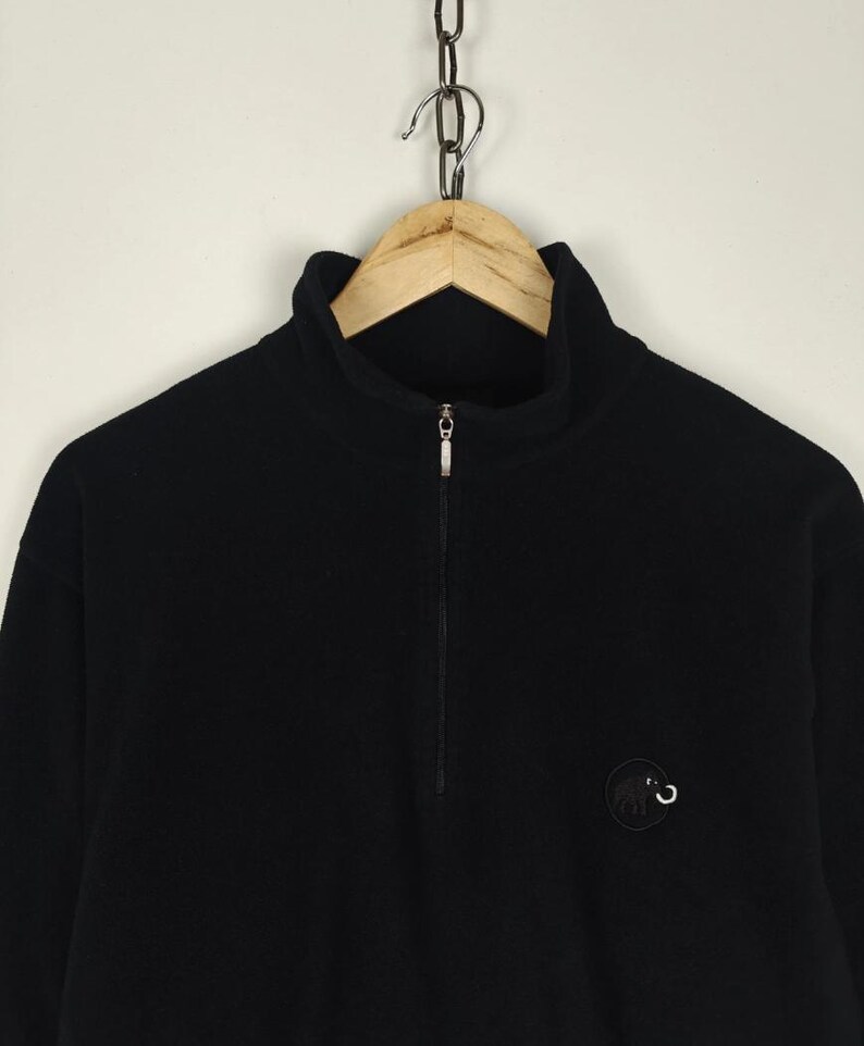 Puede incluir: Jersey polar negro con cuello alto y cremallera de un cuarto. Un peque&ntilde;o logotipo circular est&aacute; en el pecho izquierdo. La prenda cuelga de una percha de madera suspendida de una cadena de metal.