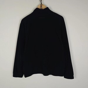 Puede incluir: Una camisa negra de manga larga con cuello alto. La prenda est&aacute; colgada de una percha de madera mediante una cadena de metal. La camisa es de color negro liso y parece estar hecha de un material suave.