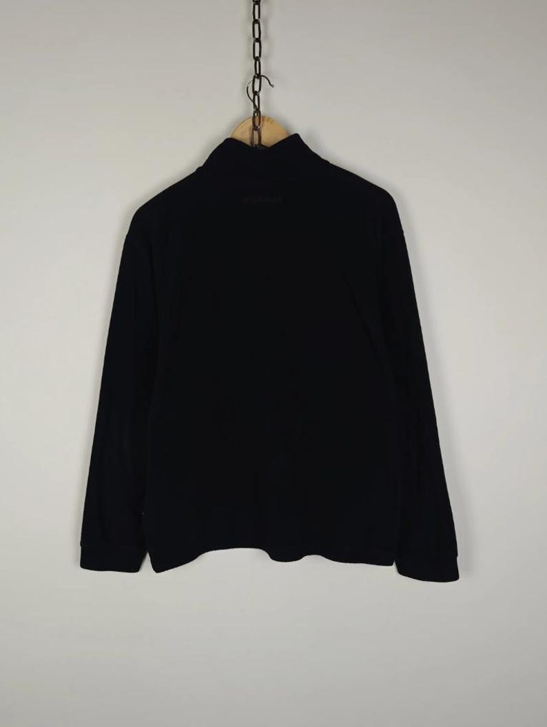 Puede incluir: Una camisa negra de manga larga con cuello alto. La prenda est&aacute; colgada de una percha de madera mediante una cadena de metal. La camisa es de color negro liso y parece estar hecha de un material suave.