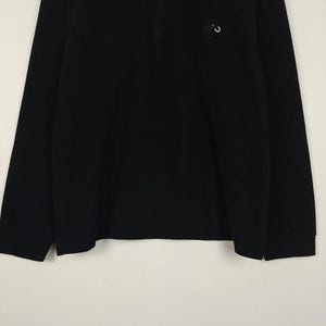 Puede incluir: Camiseta negra de manga larga con cuello con cremallera de un cuarto. Un peque&ntilde;o emblema circular est&aacute; en la parte superior izquierda del pecho. La camiseta tiene un dise&ntilde;o simple y cl&aacute;sico, adecuado para ropa casual.