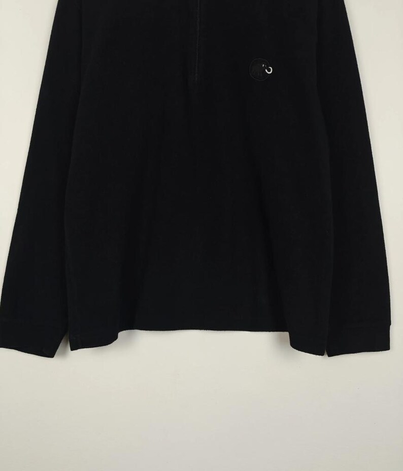 Puede incluir: Camiseta negra de manga larga con cuello con cremallera de un cuarto. Un peque&ntilde;o emblema circular est&aacute; en la parte superior izquierda del pecho. La camiseta tiene un dise&ntilde;o simple y cl&aacute;sico, adecuado para ropa casual.
