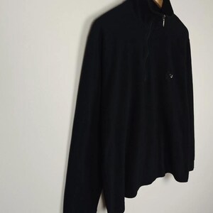 Puede incluir: Jersey negro de manga larga con cuello alzado y cierre de cremallera de un cuarto. La camisa tiene un peque&ntilde;o logotipo en el pecho y est&aacute; colgada de una percha de madera. La prenda est&aacute; hecha de un material suave y c&oacute;modo.
