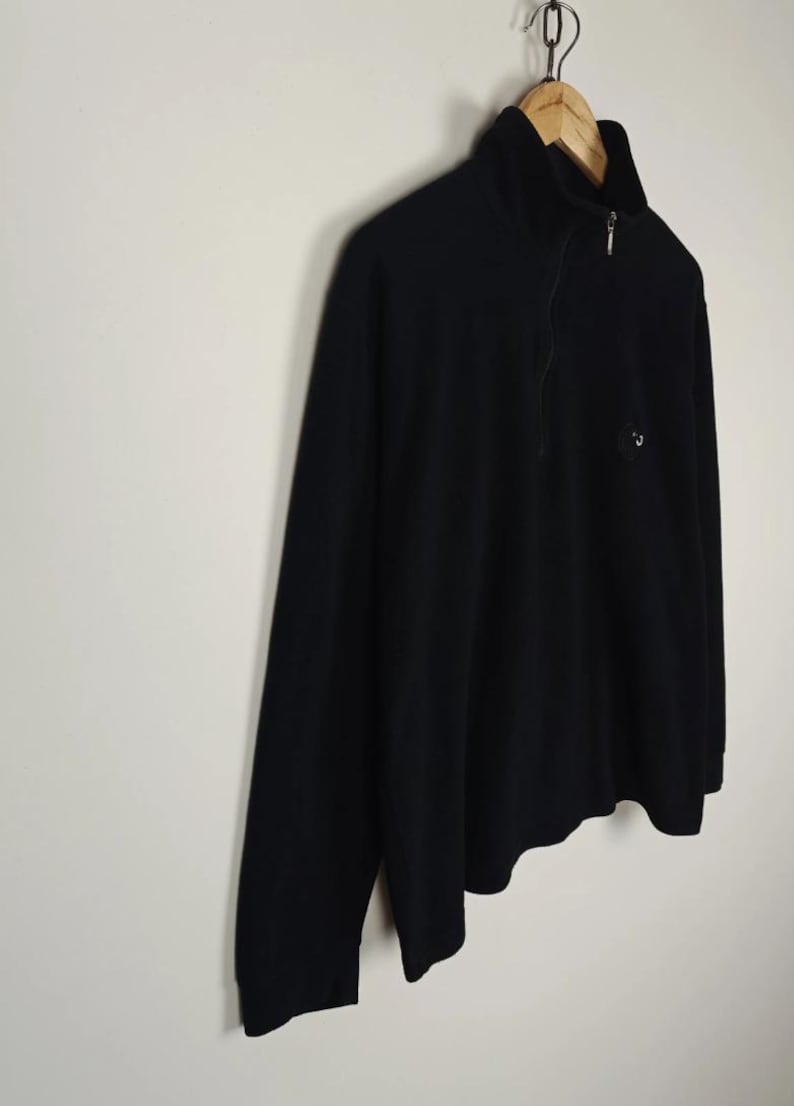 Puede incluir: Jersey negro de manga larga con cuello alzado y cierre de cremallera de un cuarto. La camisa tiene un peque&ntilde;o logotipo en el pecho y est&aacute; colgada de una percha de madera. La prenda est&aacute; hecha de un material suave y c&oacute;modo.